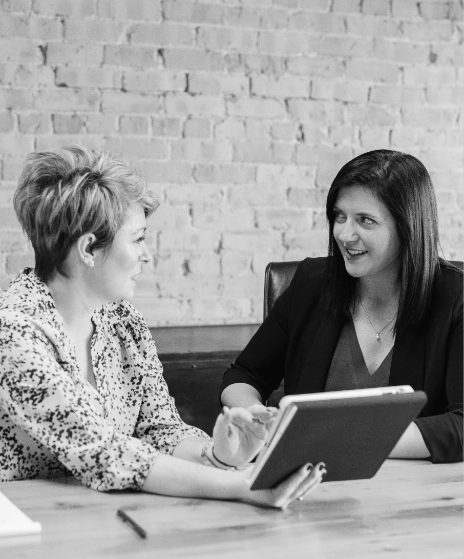 2 women sitting behind a desk talking to each other while looking at an iPad one of them is holding