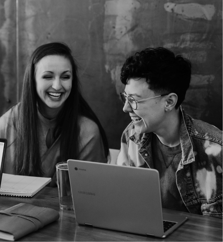 A black and white photo of two women laughing sitting at a table with laptops open in front of them