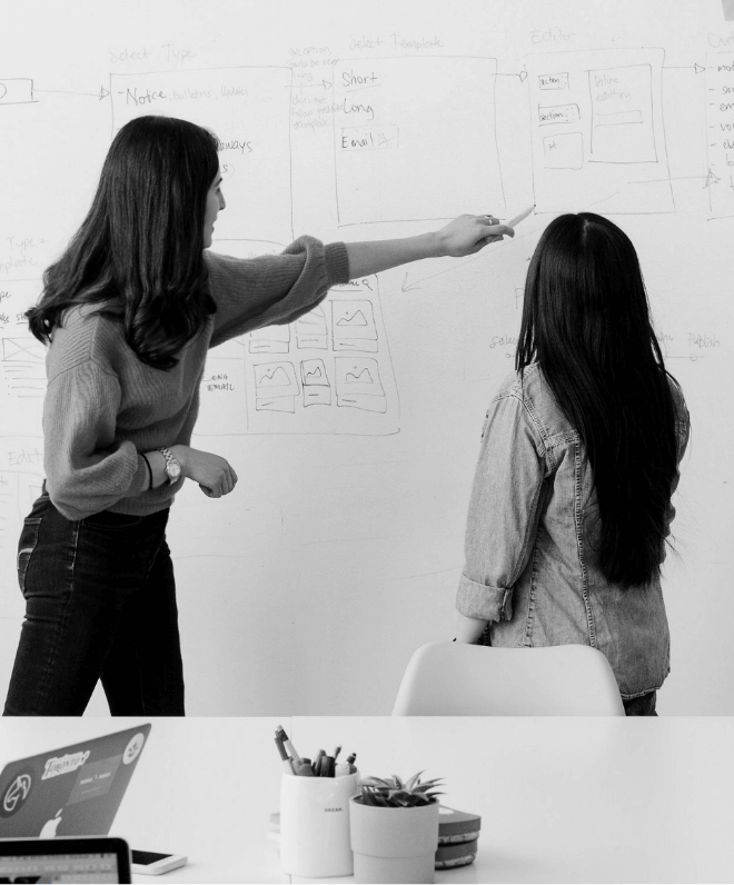 A black and white photograph of two people with their backs turned with one of them pointing to something on a whiteboard