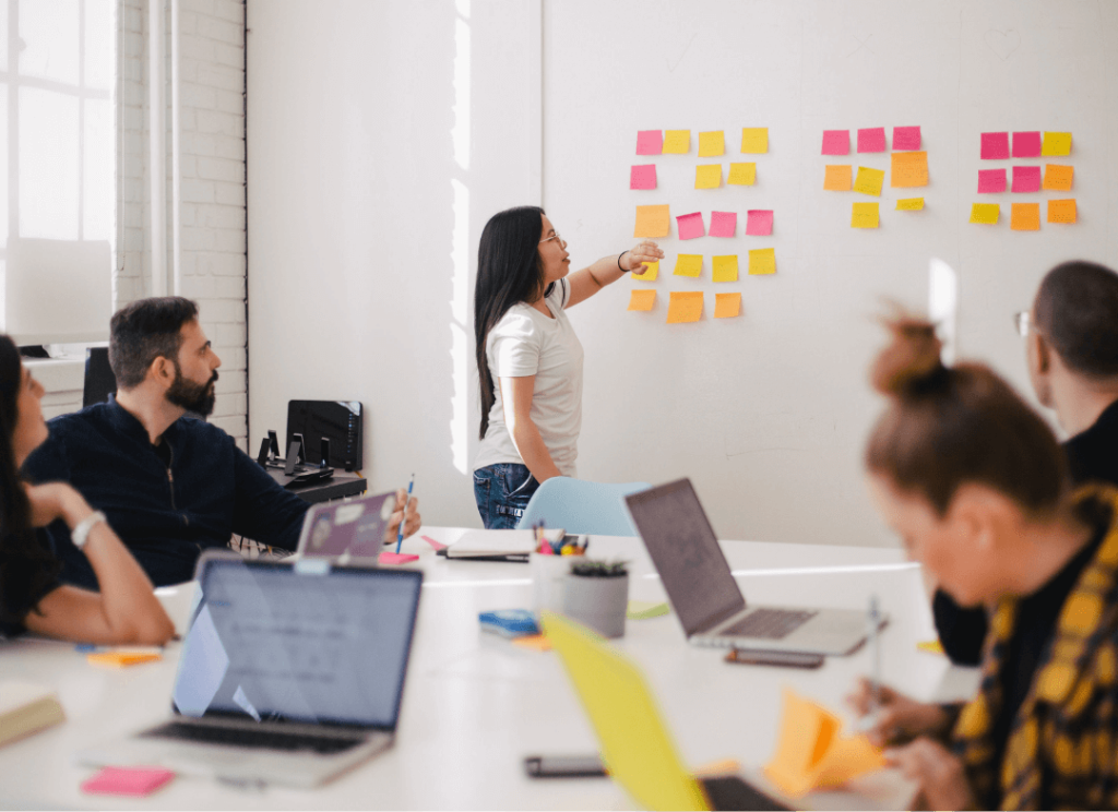 A person standing up in front of a group pointing to some post-it notes on a whiteboard